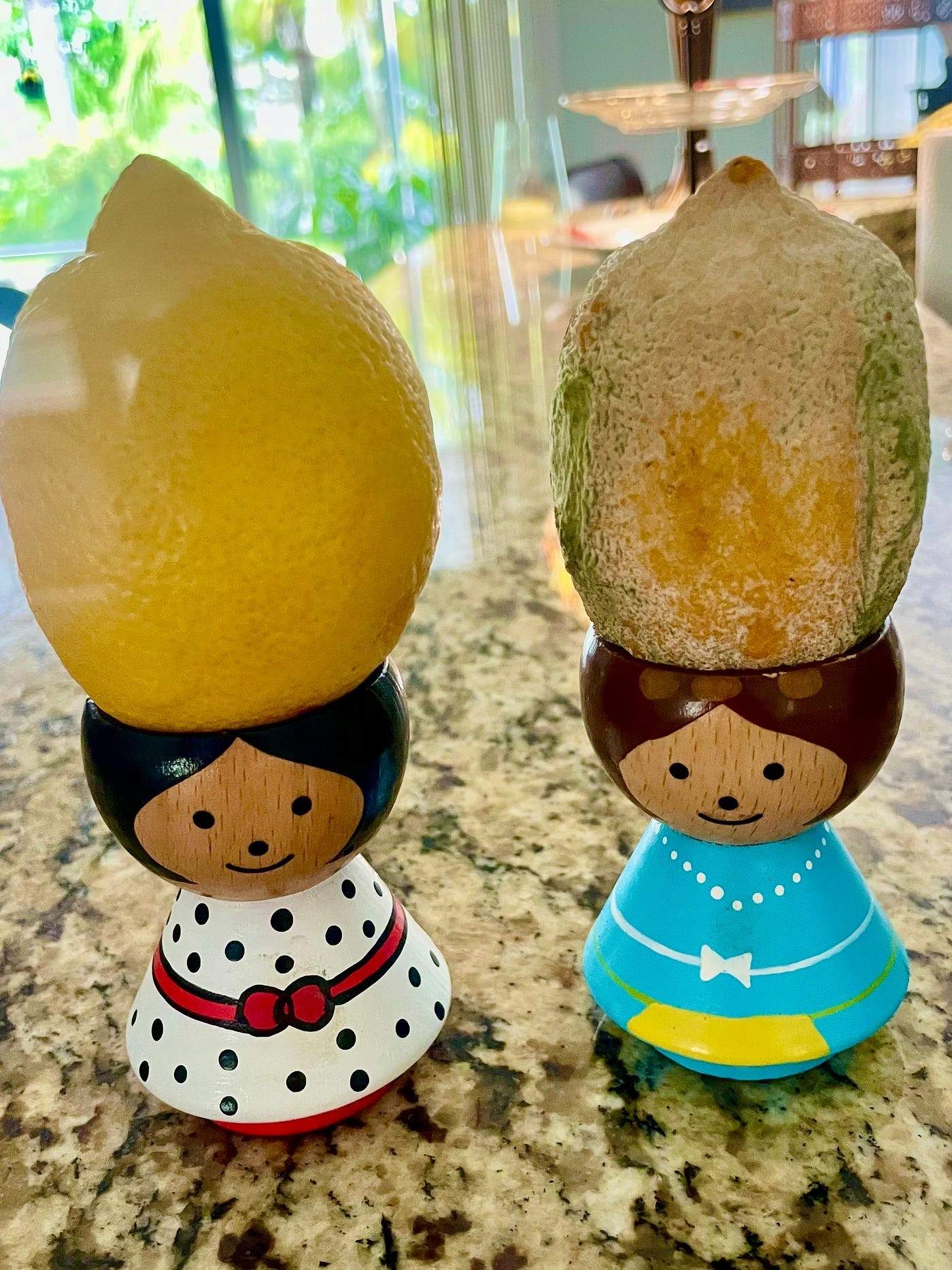 Two small wooden figurines with painted faces and dresses, each balancing a lemon on their heads